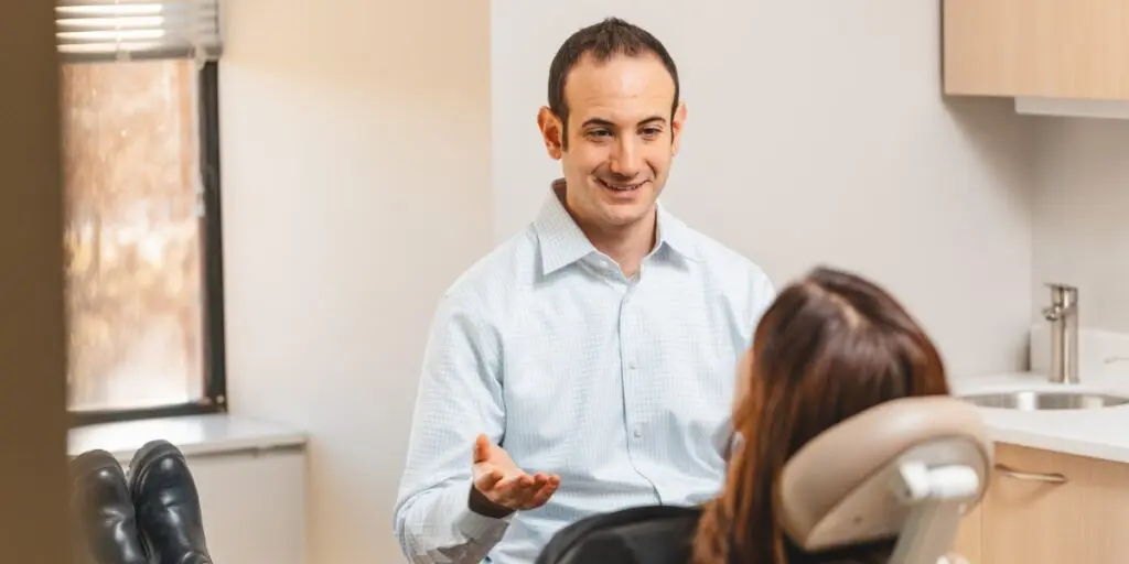 Male clinician in a light blue shirt explains something to a patient seated in a clinic chair in a bright, modern room.